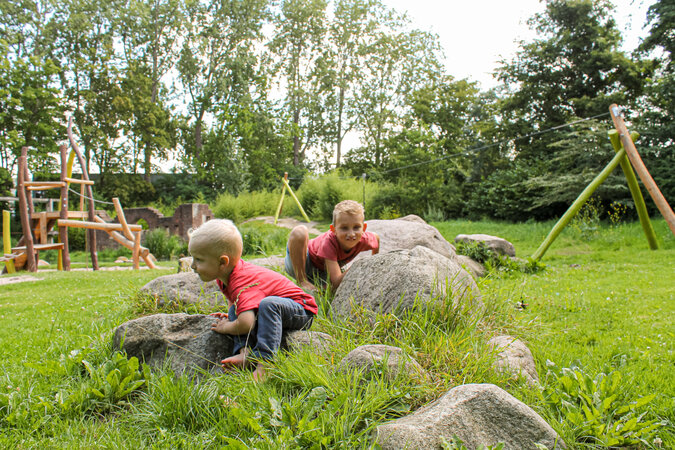 Openbare speelvoorzieningen - twee jongens spelen tussen grote stenen in het groen vóór een eibe speeltuin.