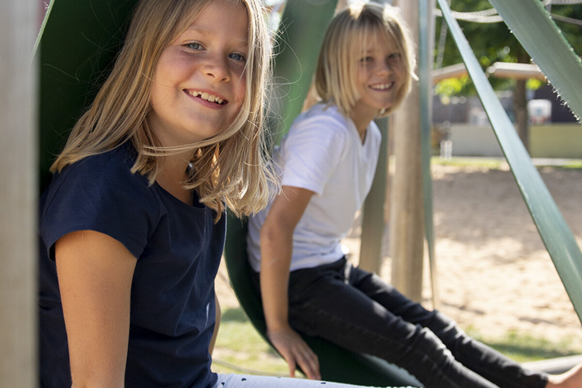 Schoolspeelplaats planning - Twee lachende meisjes zitten in eibe speeltoestellen op hun schoolspeelplaats.